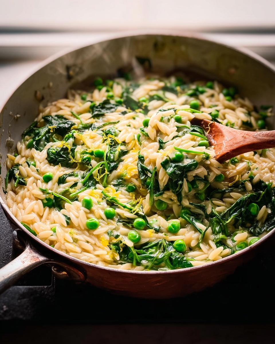 Close-up of a pan of 20-minute orzo with peas and spinach, being stirred with a wooden spoon.