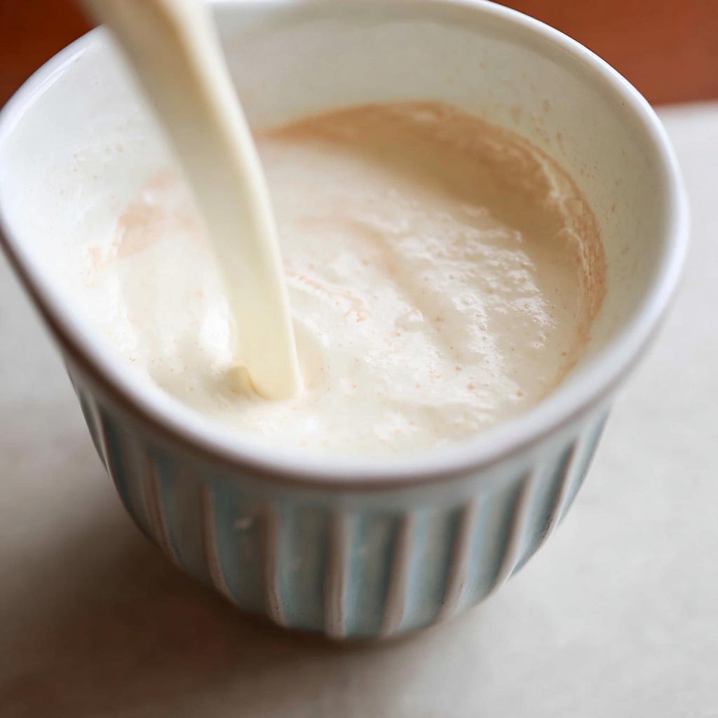 A stream of white liquid being poured into a light blue ramekin containing a creamy mixture for Champagne Cream Sauce for Seafood.