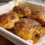Four perfectly baked chicken thighs with a crispy, seasoned crust in a white baking dish.