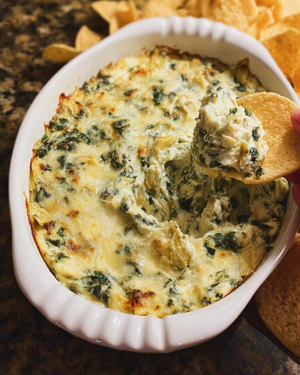 A scoop of creamy, cheesy Baked Spinach Artichoke Dip being lifted out of a white dish with a tortilla chip.