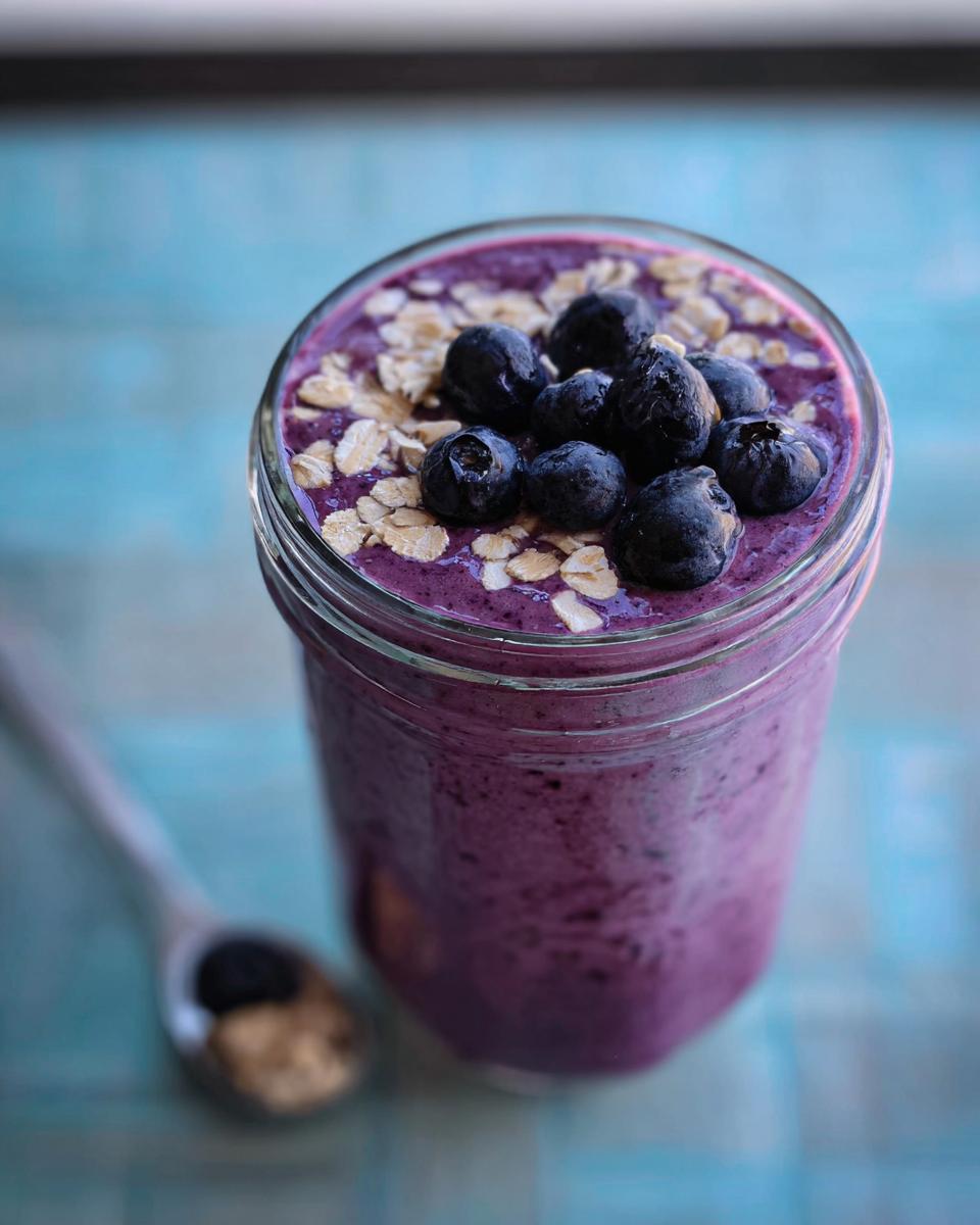 Close-up of a thick Blueberry Oat Breakfast Smoothie in a glass jar, topped with fresh blueberries and rolled oats.