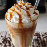 A tall glass of a blended Caramel Frappuccino (Copycat) topped high with whipped cream and drizzled caramel sauce.