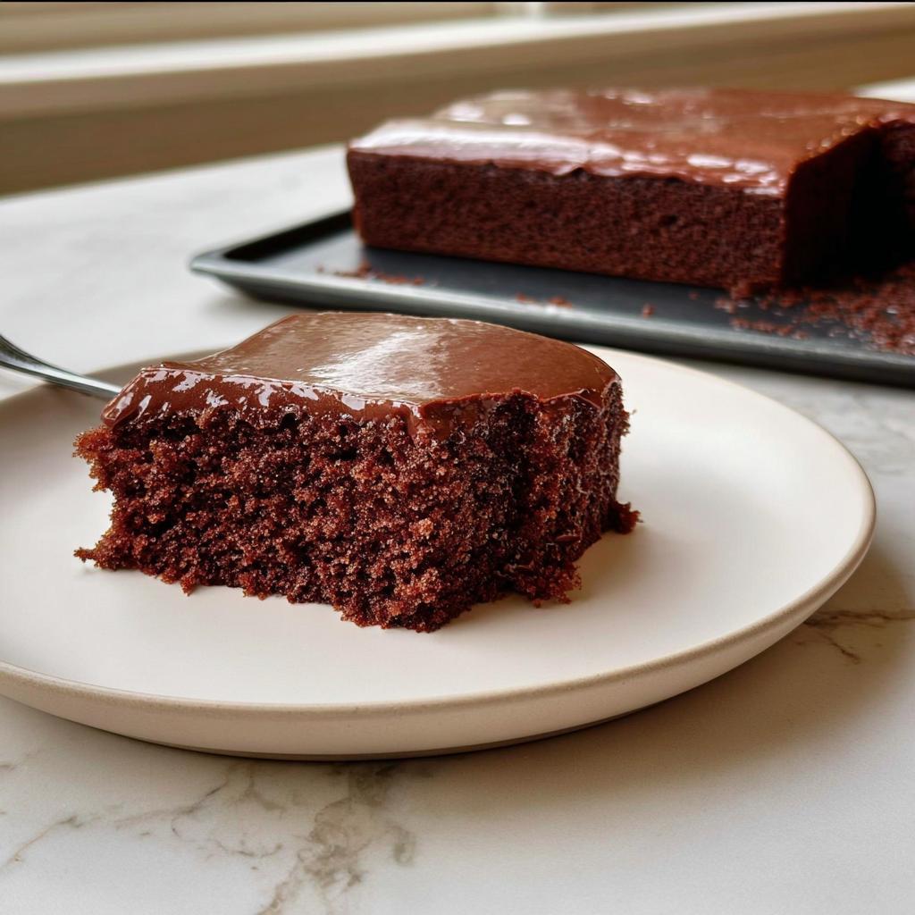 A moist slice of Easy Snacking Sheet Cake topped with shiny chocolate frosting on a white plate.