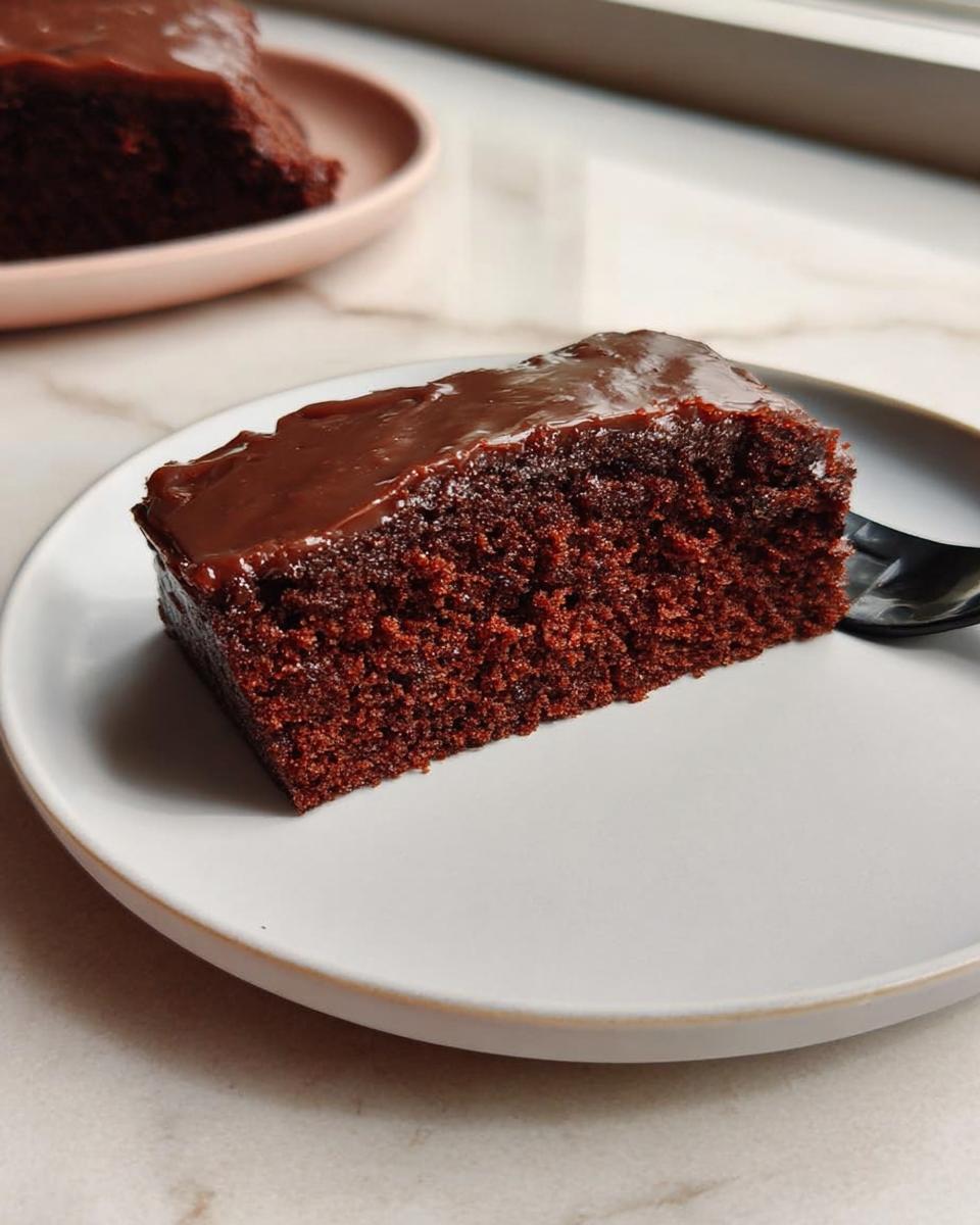 A moist slice of Easy Snacking Sheet Cake topped with glossy chocolate frosting on a white plate.