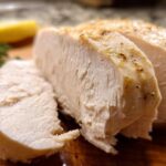 Close-up of a perfectly cooked, juicy chicken breast, sliced to reveal tender white meat. A lemon wedge and herb sprig are visible.