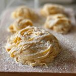Close-up of nests of fresh, homemade pasta dusted with flour on a wooden board, ready for pasta recipes.