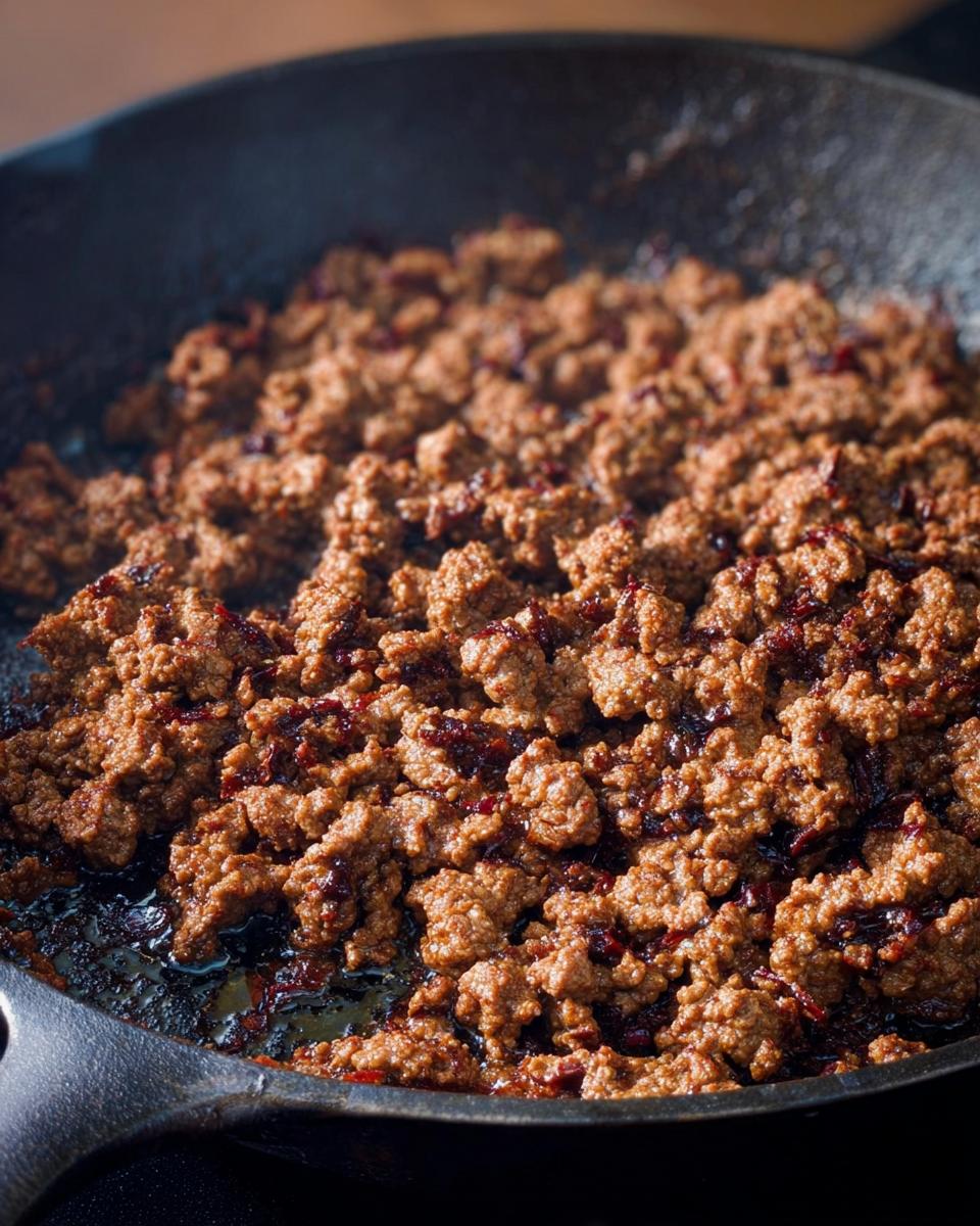 Close-up of ground beef browning in a cast iron skillet, with small dark red bits mixed in.