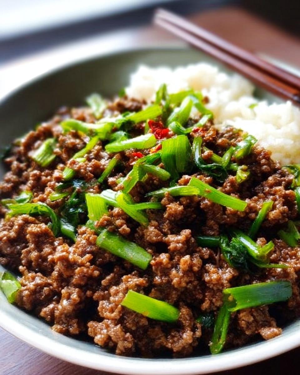 A close-up of a bowl of ground beef stir-fry with green onions and chili flakes, served with white rice. A quick and easy ground beef recipe.
