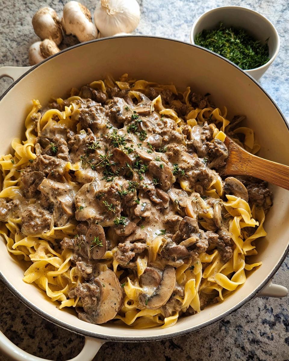A large pot filled with creamy Ground Beef Stroganoff served over wide egg noodles, garnished with fresh thyme.