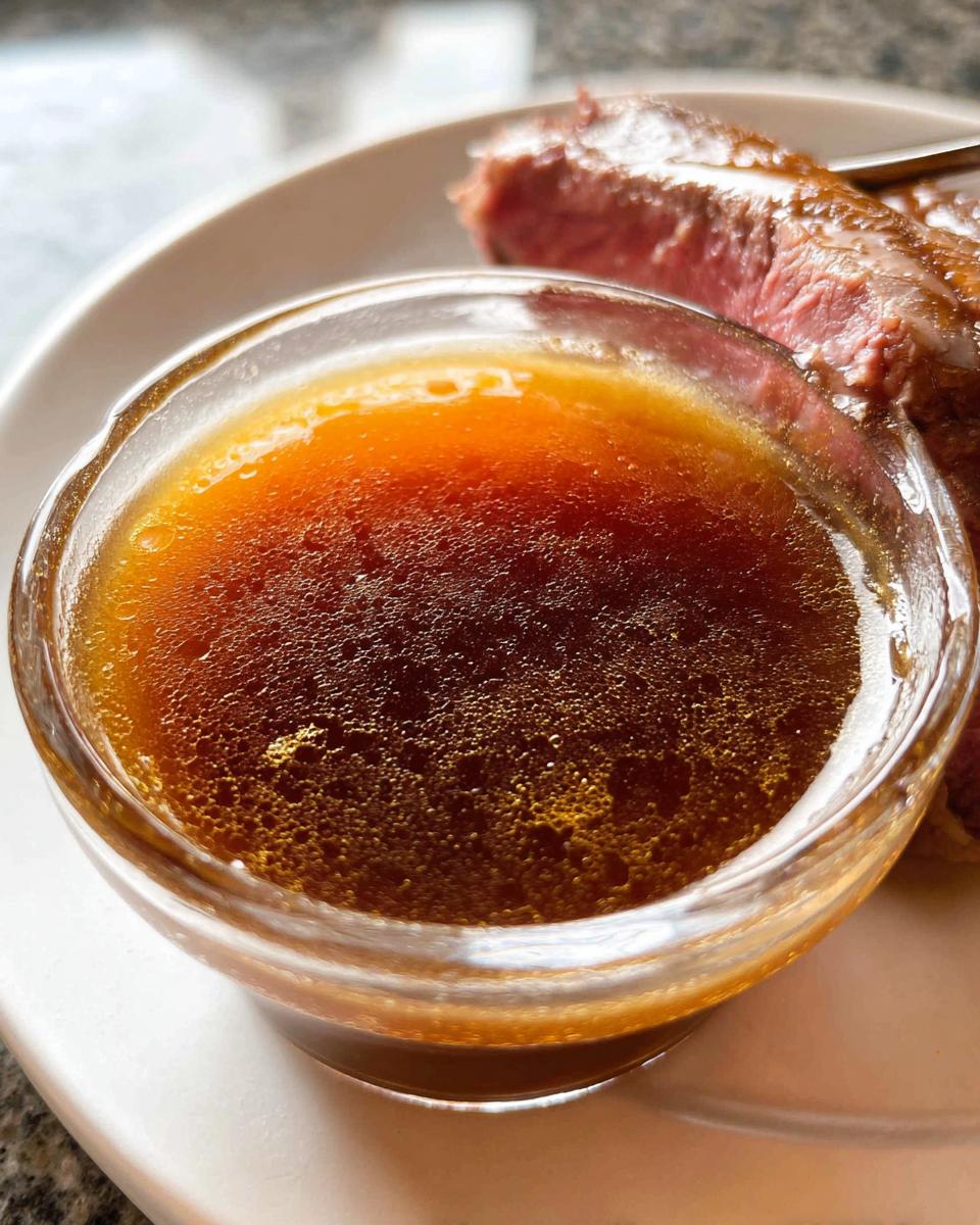 Close-up of rich, dark brown Homemade Au Jus without drippings served in a small glass bowl next to sliced roast beef.