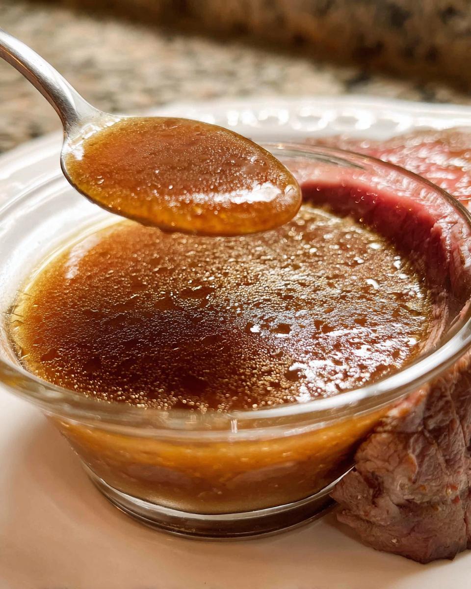 A spoonful of rich, brown Homemade Au Jus without drippings being lifted from a small glass bowl next to a slice of roast beef.