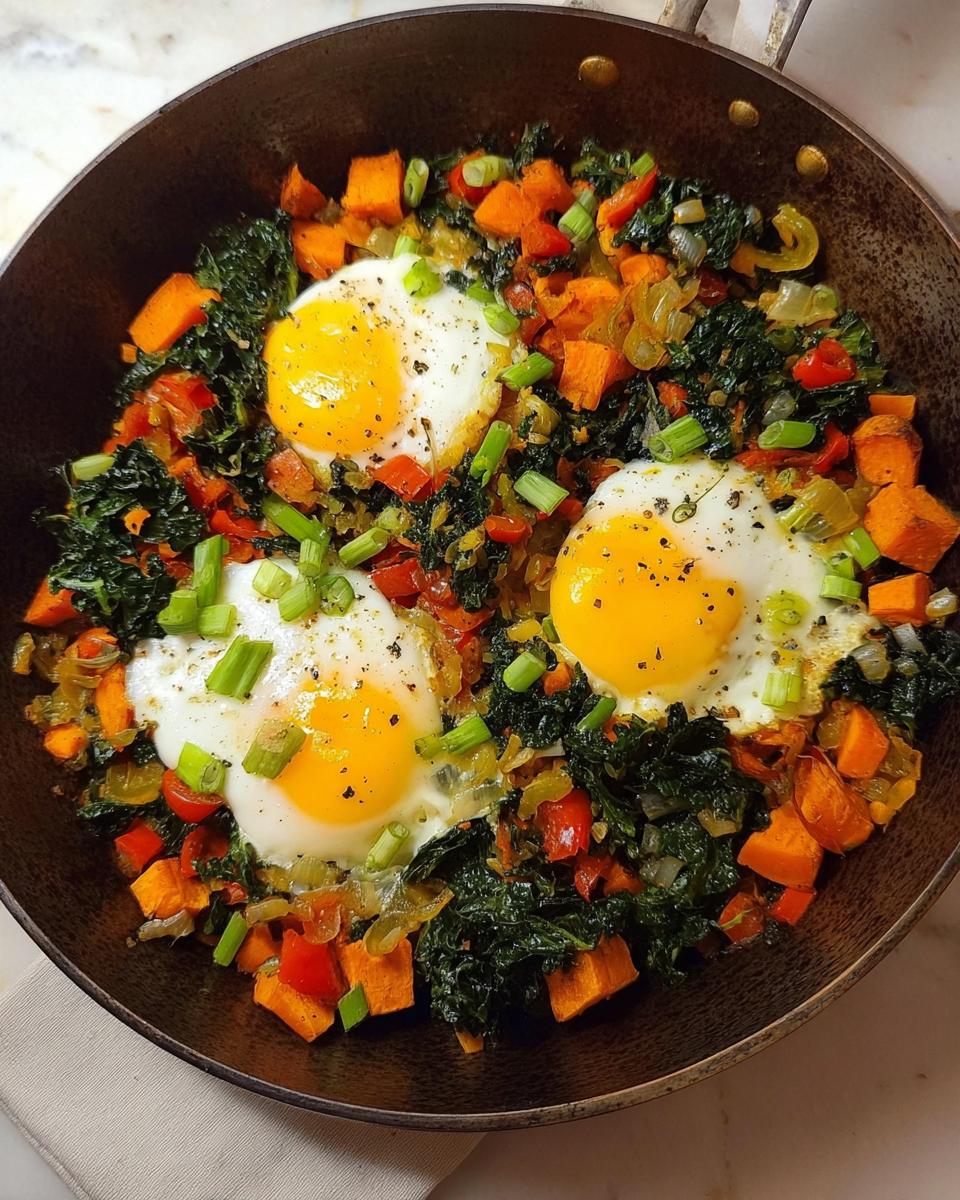 A close-up of a one-pan breakfast with three sunny-side-up eggs nestled in a medley of kale, diced sweet potatoes, and red peppers, topped with green onions.