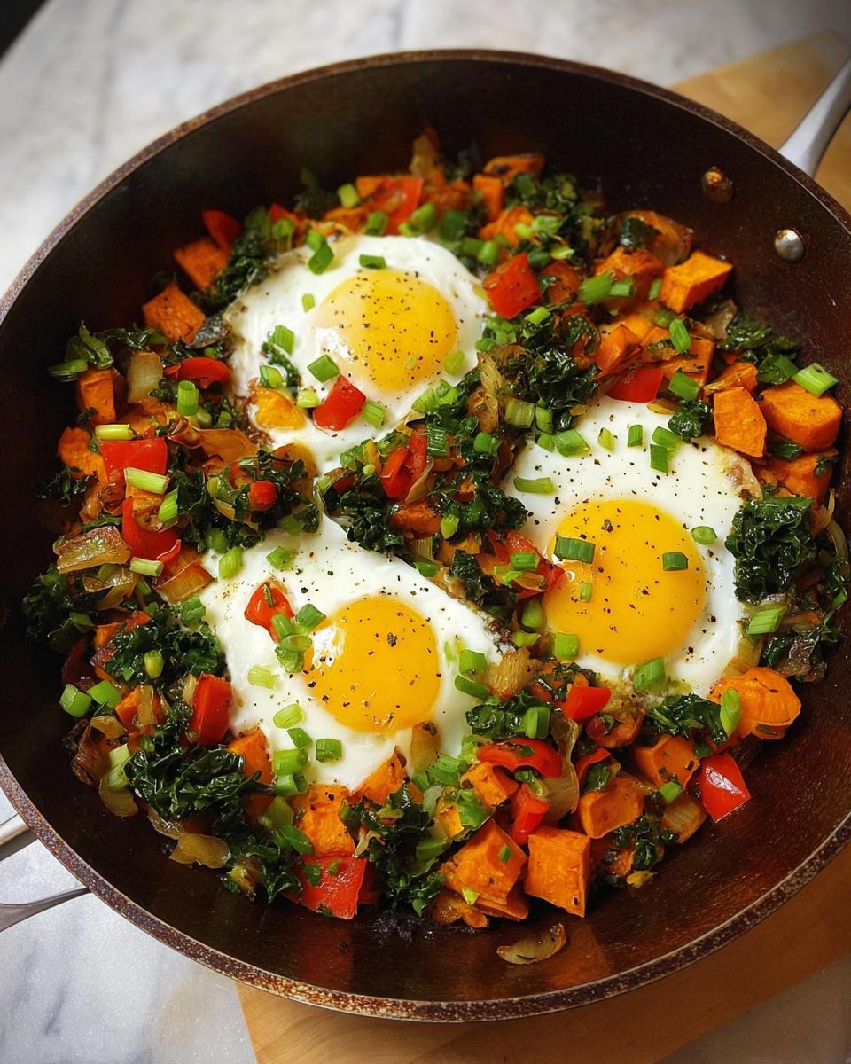 A close-up of a one-pan breakfast with fried eggs nestled in a colorful mix of sweet potatoes, bell peppers, kale, and onions.