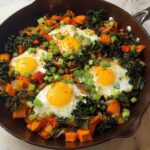 A hearty one-pan breakfast featuring fried eggs nestled in a colorful hash of sweet potatoes, kale, onions, and peppers, sprinkled with chives.