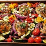 A close-up of a sheet pan filled with golden-brown One-Pan Chicken pieces, colorful roasted vegetables like zucchini, bell peppers, red onion, tomatoes, and olives, topped with feta cheese and herbs.