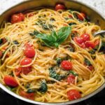 A close-up of a one-pan pasta recipe with spaghetti, cherry tomatoes, spinach, and basil.