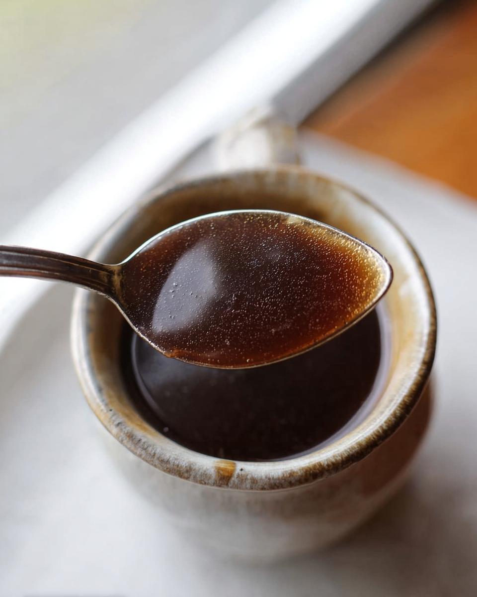 Close-up of a spoonful of rich, dark brown Au Jus Recipe liquid being lifted from a small ceramic cup.