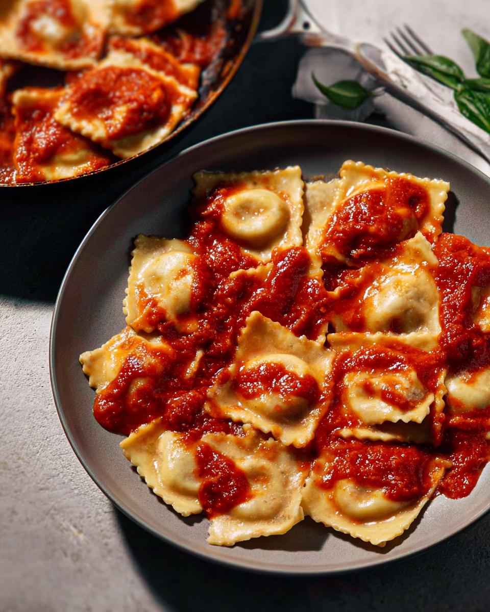 Close-up of a plate of ravioli covered in marinara sauce, with more ravioli in a pan in the background.