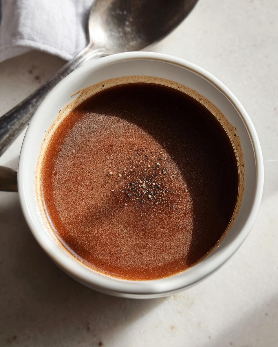 Close-up of rich, dark brown au jus topped with cracked pepper in a white mug.