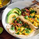 Two scrambled egg breakfast tacos filled with pico de gallo and cilantro, served with sliced avocado and lime. Part of Ultimate Breakfast Ideas.