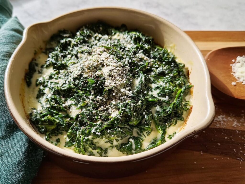 A close-up of rich, dark green Creamed Spinach (Steakhouse Style) topped with grated Parmesan and black pepper in a beige baking dish.
