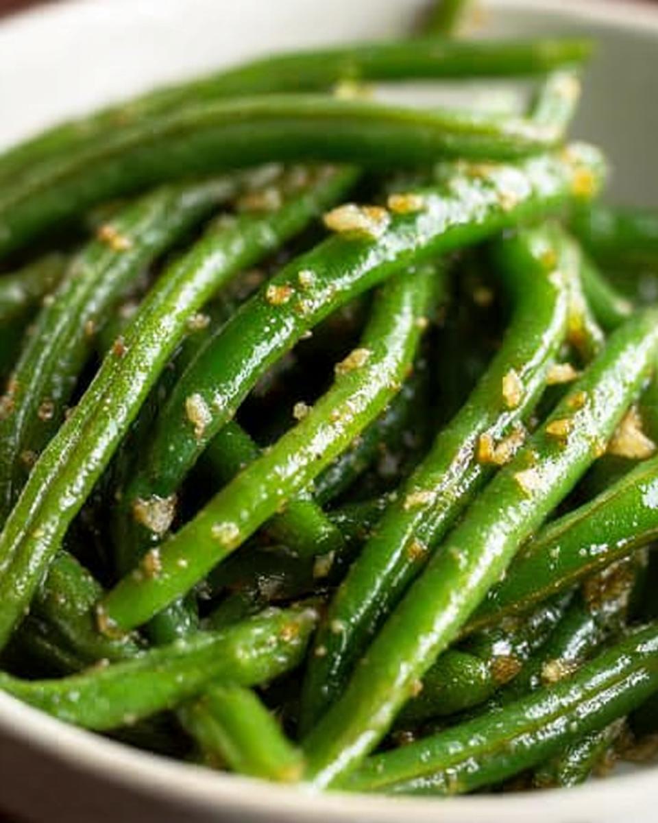 Close-up of glossy Thanksgiving green beans tossed with minced garlic and seasoning in a white bowl.