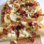 Close-up of a slice of 10-ingredient pizza featuring pear, crispy bacon, and crumbled blue cheese on a white serving board.