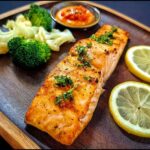 A perfectly cooked salmon fillet seasoned with herbs, served with steamed broccoli, cauliflower, lemon slices, and a dipping sauce.