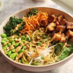 A vibrant and healthy 20-ingredient rice bowl filled with edamame, tofu, shredded carrots, and greens, drizzled with a creamy sauce.