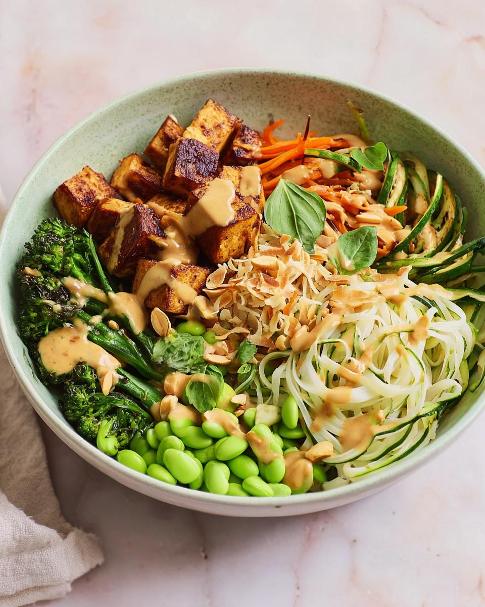 A vibrant bowl of 20-Ingredient Rice Bowls with tofu, edamame, zucchini noodles, and broccoli, drizzled with peanut sauce.