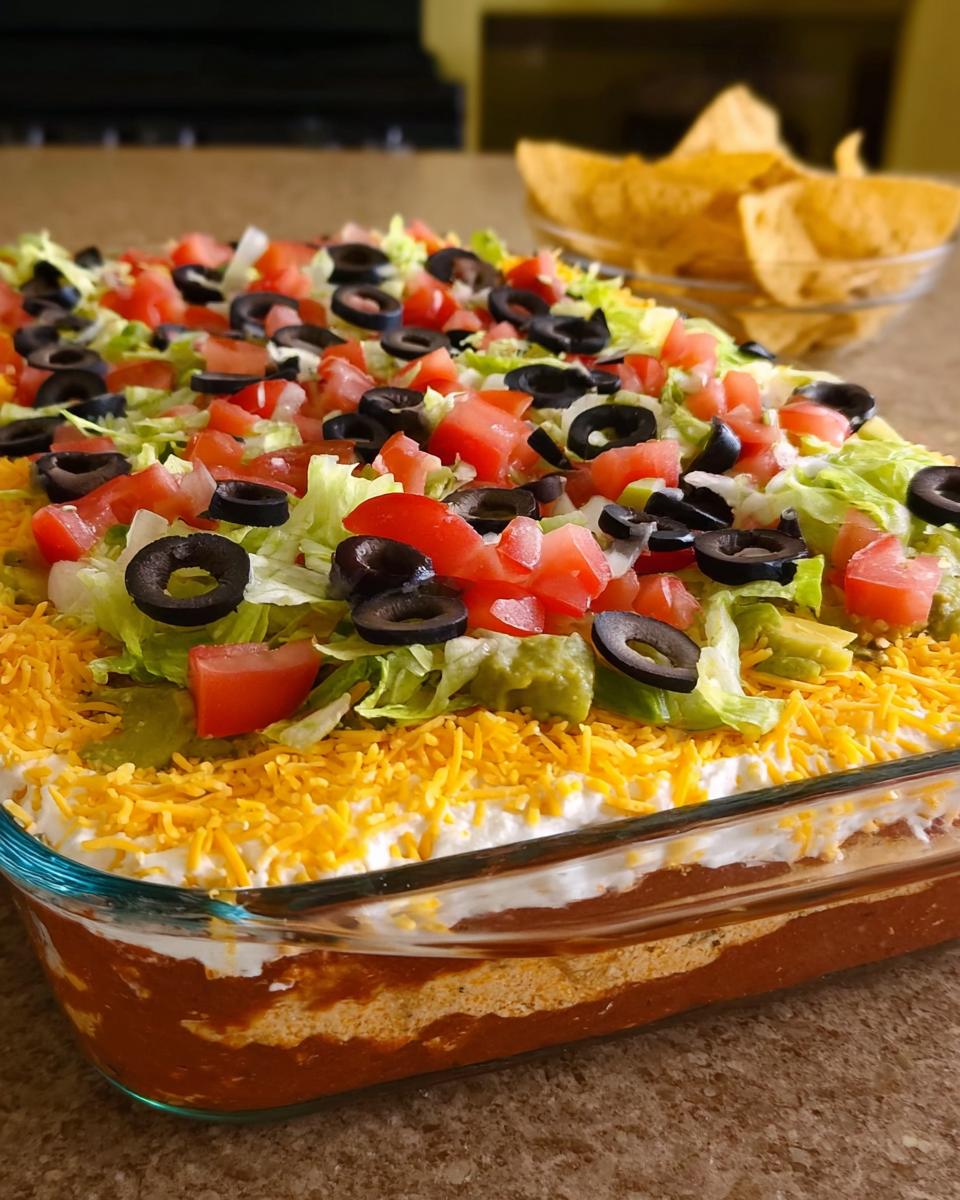 A delicious 7 layer taco dip, perfect for Taco Tuesday recipes, topped with lettuce, tomatoes, olives, and cheese.
