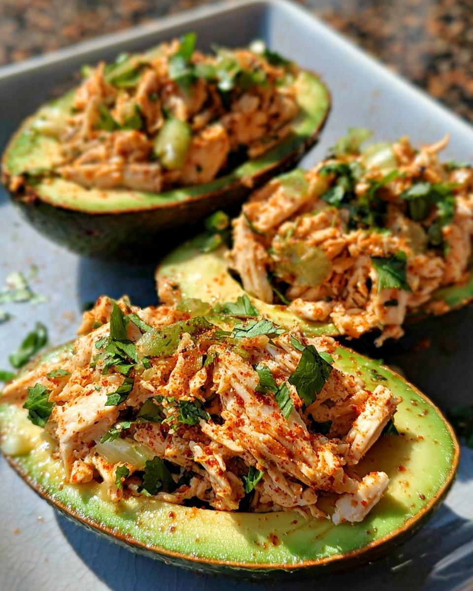 Three halves of avocado stuffed with shredded chicken salad, garnished with fresh herbs and paprika. A quick sandwich idea.