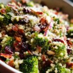A close-up of a vibrant broccoli and quinoa salad featuring crispy bacon, dried cranberries, slivered almonds, and grated Parmesan cheese.