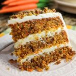A slice of moist carrot cake with cream cheese frosting and chopped nuts, showcasing layers of cake and frosting.
