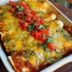 Close-up of baked chicken enchiladas in a white dish, topped with melted cheese, salsa verde, and diced tomatoes.