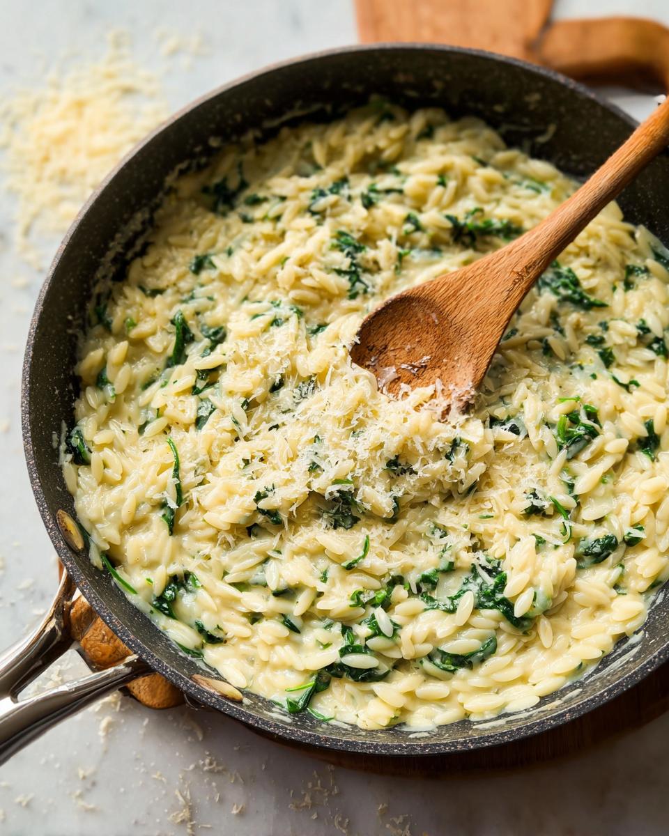 Close-up of creamy orzo pasta with spinach and grated cheese in a pan, mixed with a wooden spoon. A perfect veggie sides recipe.