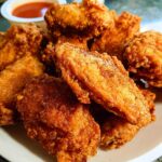 A plate of golden-brown, crispy fried chicken wings, seasoned with spices, served with a side of dipping sauce.