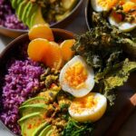 A vibrant bowl featuring a hard-boiled egg recipe, purple rice, sliced avocado, and orange segments.