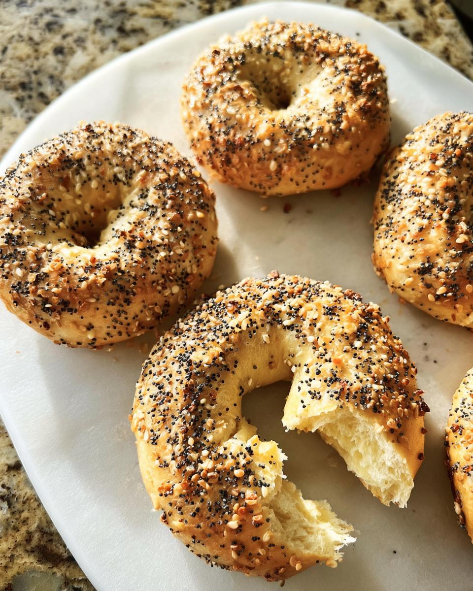 A close-up of several everything bagels, one with a bite taken out, showcasing the poppy and sesame seed topping.