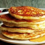 A stack of fluffy pancakes drizzled with syrup, with a bowl of berries in the background. A perfect breakfast idea.