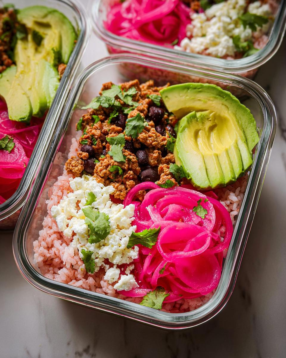 Overhead view of a meal prep rice bowl recipe with pink rice, seasoned ground turkey, black beans, crumbled feta, pickled red onions, and avocado slices.