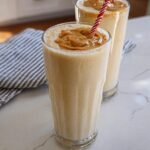 Two tall glasses filled with creamy peanut butter smoothies, topped with a dollop of peanut butter and a striped straw. A perfect beginner breakfast idea.