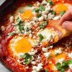A hand dipping bread into a skillet of restaurant-style eggs, cooked in a rich tomato sauce and topped with feta cheese and cilantro.