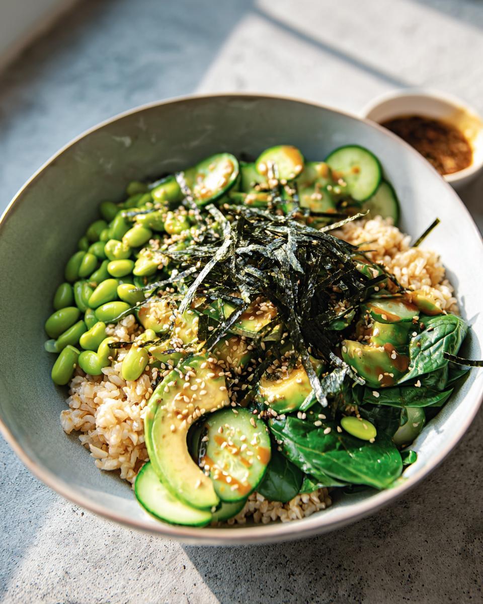 A vibrant and healthy rice bowl featuring brown rice, sliced avocado, edamame, cucumber, spinach, and nori, drizzled with sauce.