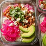 Overhead view of a meal prep container filled with rice bowls recipes: white rice, pink rice, seasoned ground meat, crumbled feta, pickled red onions, cilantro, and avocado slices.