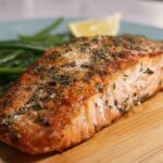 A close-up of a perfectly cooked salmon fillet, seasoned with herbs, served with steamed green beans and a lemon wedge.