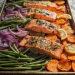 Four fillets of salmon seasoned and baked on a sheet pan with sliced sweet potatoes, green beans, red onions, and lemon wedges.