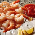 A close-up of a shrimp cocktail appetizer, featuring cooked shrimp in a martini glass with cocktail sauce and lemon wedges.