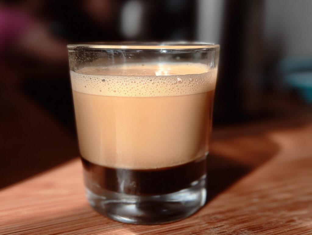 A layered Starbucks drink recipe idea in a shot glass, featuring dark espresso at the bottom and creamy frothed milk on top.