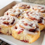 Close-up of fluffy strawberry breakfast rolls drizzled with white icing in a baking dish.